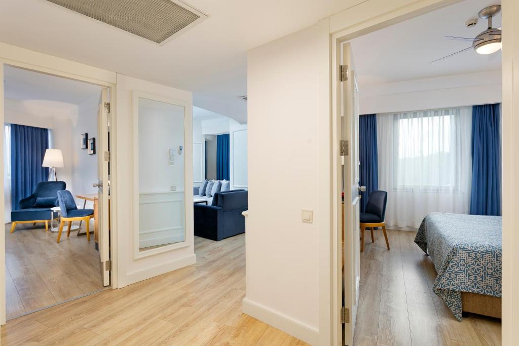 Mūsdienīgs viesnīcas numuru interjers ar divām durvīm, kas ved uz viesistabu un guļamistabu, ar koka grīdu un gaišiem, moderniem mēbeļu elementiem.
