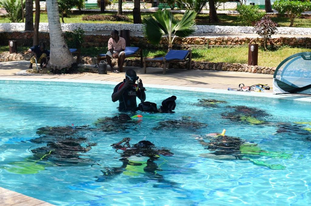 Peldēšanas nodarbība baseinā, kur instruktors vada grupu ar snorkelēšanas aprīkojumu. Apkārt ir koki un atpūtas zonas, radot relaksējošu atmosfēru.