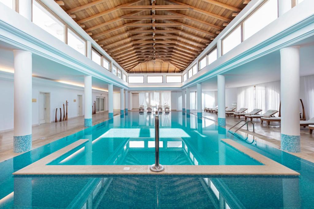 Mūsdienīgs un elegants spa baseins ar plašām logiem un koka jumtu, kas nodrošina gaišu un relaksējošu atmosfēru. Ūdens ir dzidrs un aicinošs, blakus ir atpūtas krēsli.