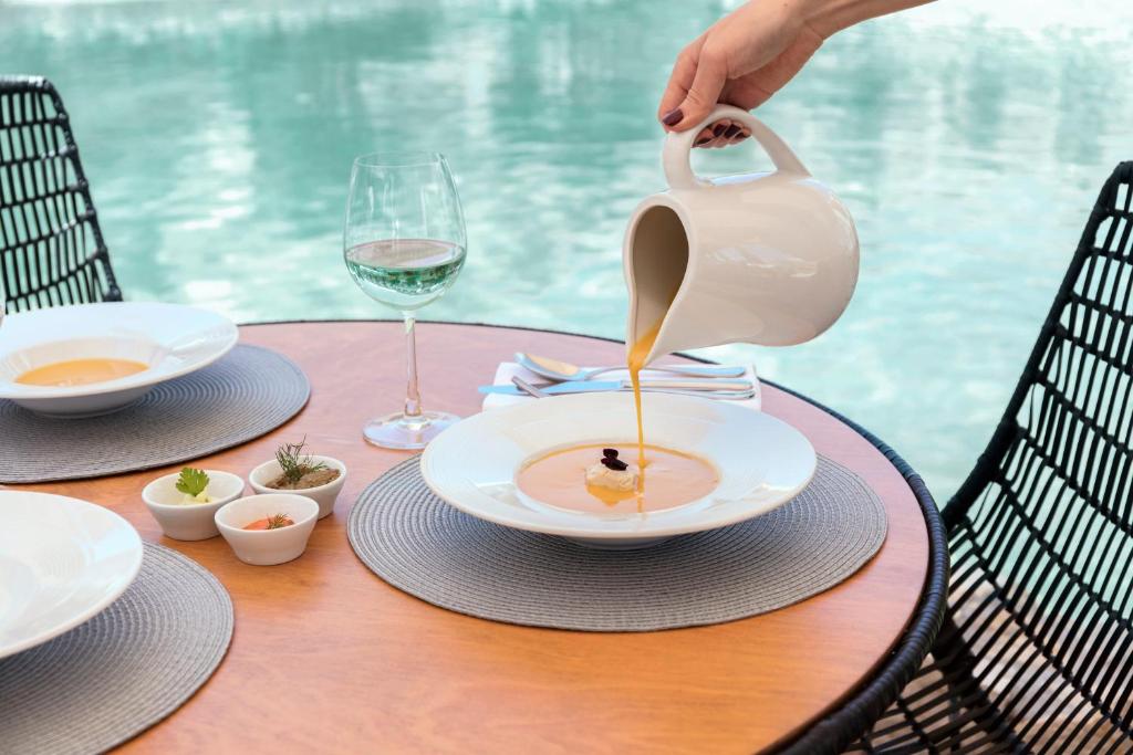 Eleganta maltīte pie galda ar skatu uz ūdeni, kur kāds lej zupu no balta krūka. Uz galda ir divas zupas šķīvji un vīna glāze, kā arī mazi trauki ar garšvielām.