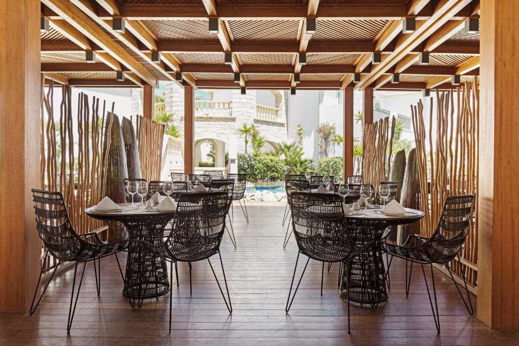 Moderns restorāns ar elegantām melnām vītņu krēslām un galdiem, kas izvietoti zem koka nojumes. Attēlā redzama zaļa ainava un peldbaseins, kas papildina atmosfēru.