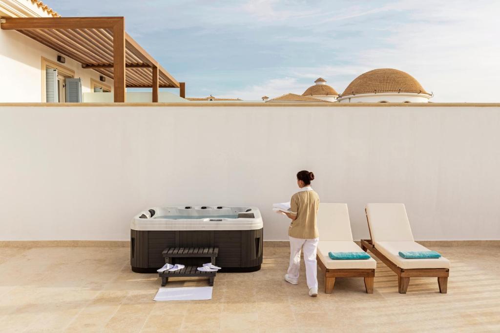 Relaksējoša ainava ar džakuzi un divām sauļošanās krēslām, kurā redzama darbiniece, kas sagatavo teritoriju. Fons ar modernu arhitektūru un skaidru debesu ainavu.