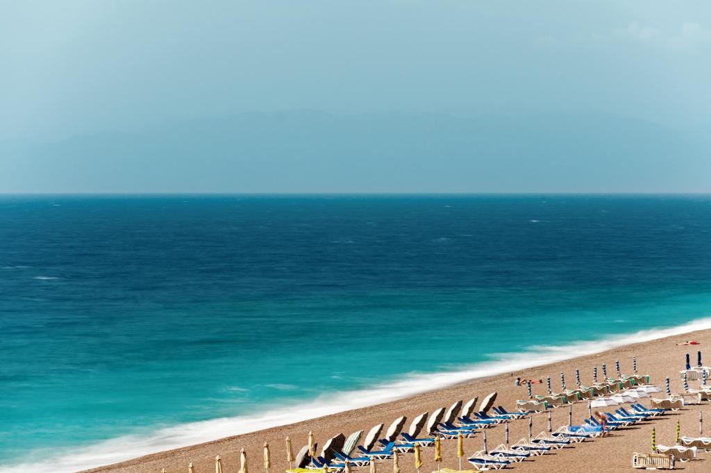 Pludmales skats ar saulessargiem un sauļošanās krēsliem, kas izvietoti pie zilas jūras un smilšu krasta. Jūra pārvēršas no tumši zilas uz gaišāku toni, radot mierīgu atmosfēru.