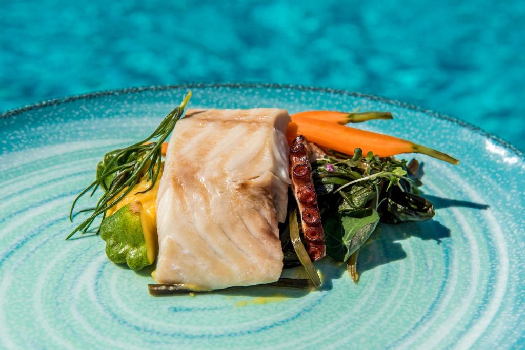 Gards zivju ēdiens ar dārzeņiem, pasniegts uz krāsainas šķīvja, fona ir zila ūdens virsma.