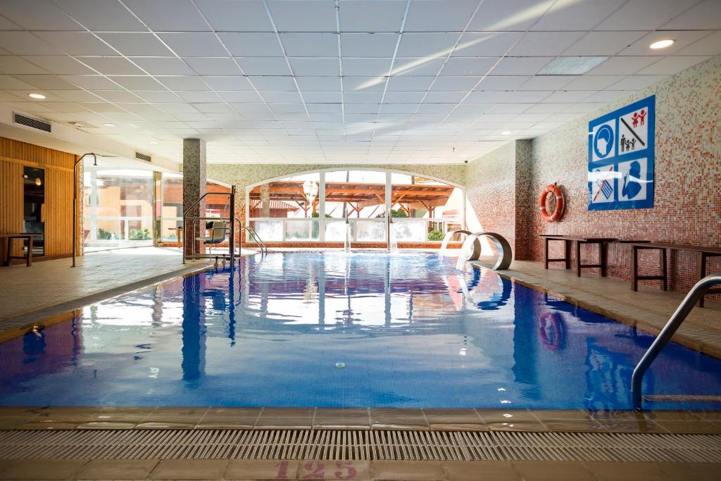 Iekštelpu baseins ar stikla sienām, kas nodrošina dabīgu apgaismojumu un modernu dizainu. Apkārt baseinam ir koka mēbeles un drošības simboli uz sienas.