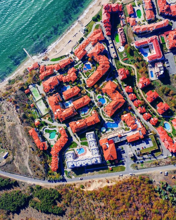 Gaisa skats uz viesnīcu kompleksu pie jūras, kur redzamas sarkanas jumtu ēkas, peldbaseini un zaļās zonas. Apburošs ainavas skats, kas atspoguļo gan dabu, gan cilvēku radītās struktūras.