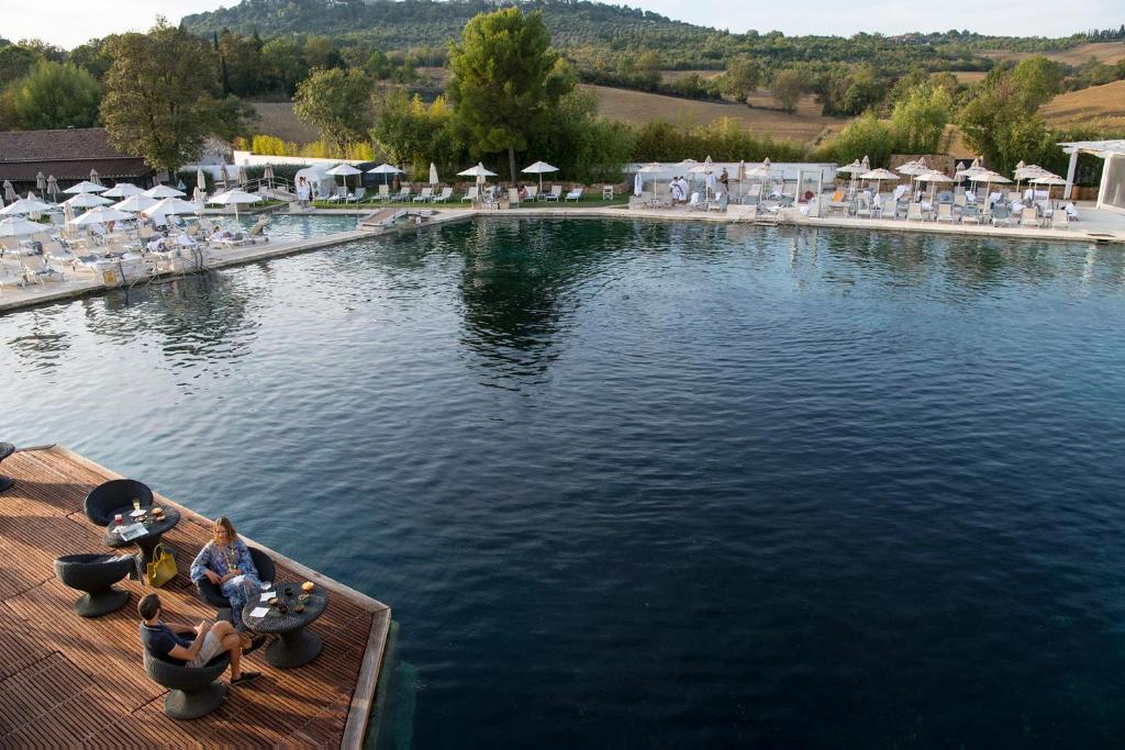 Elegants baseins ar saulessargiem un atpūtas zonām, kur divas personas sarunājas uz koka terases, baudot mierīgu atmosfēru un skaisto ainavu.