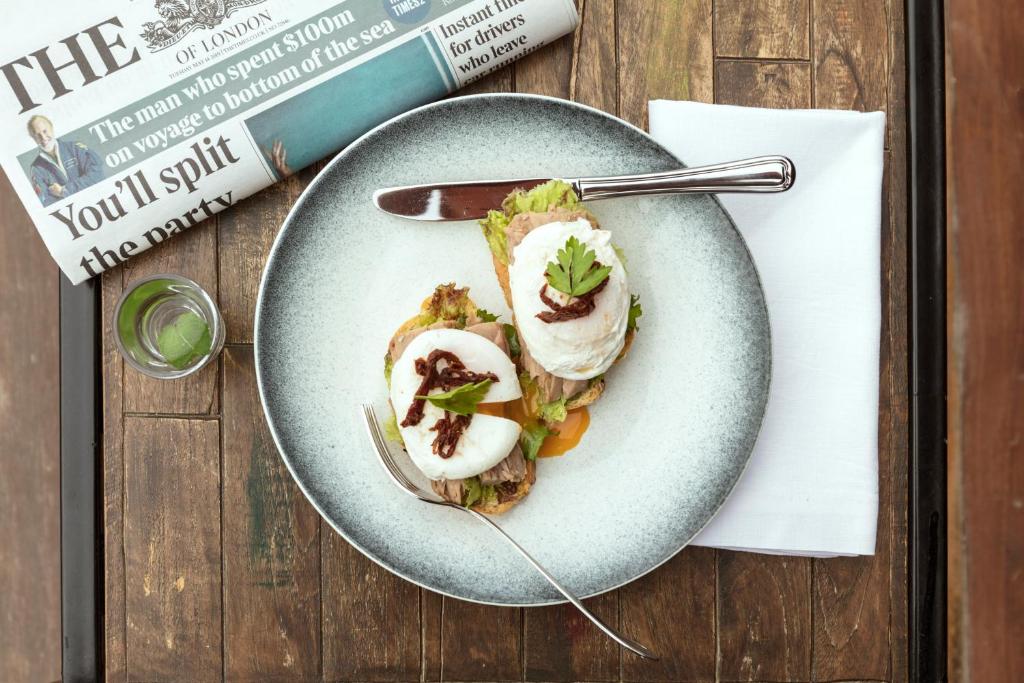 Svaigā brokastu plate ar pošētiem olām, avokado un žāvētiem tomātiem, blakus glāze ar ūdeni un laikraksts uz koka galda.