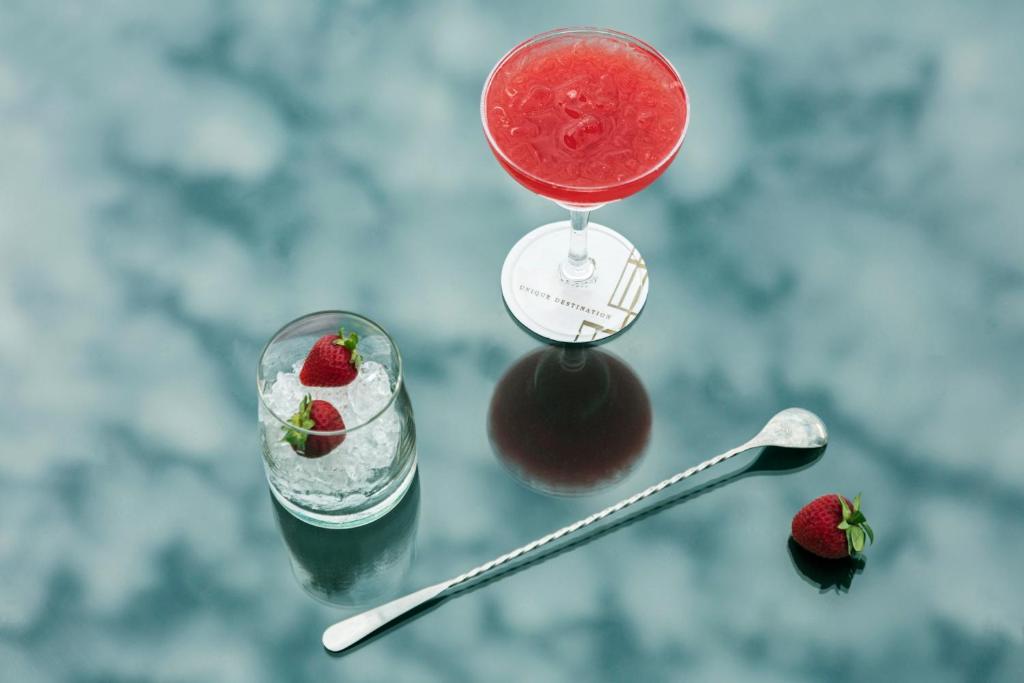 Sarkans kokteilis ar zemenēm, kas pasniegts elegantā glāzē, blakus atrodas šķīvis ar ledus un zemenēm, kā arī kokteiļu karote.