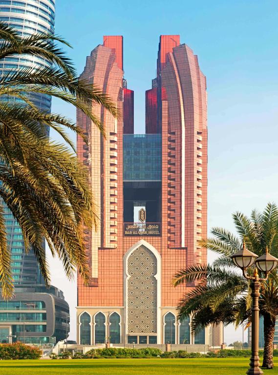 Mūsdienīga arhitektūra ar sarkanu fasādi un tradicionāliem elementiem, redzama Bab Al Qasr viesnīca, kas atrodas starp palmām un modernām ēkām.