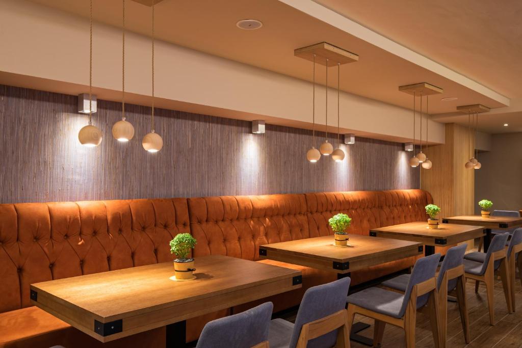 Mūsdienīgs restorāna interjers ar brūnu ādas dīvānu, koka galdiem un pakarināmām lampām, kas rada siltu atmosfēru. Uz galdiem ir nelieli zālītes podi, kas piešķir dzīvīgumu telpai.