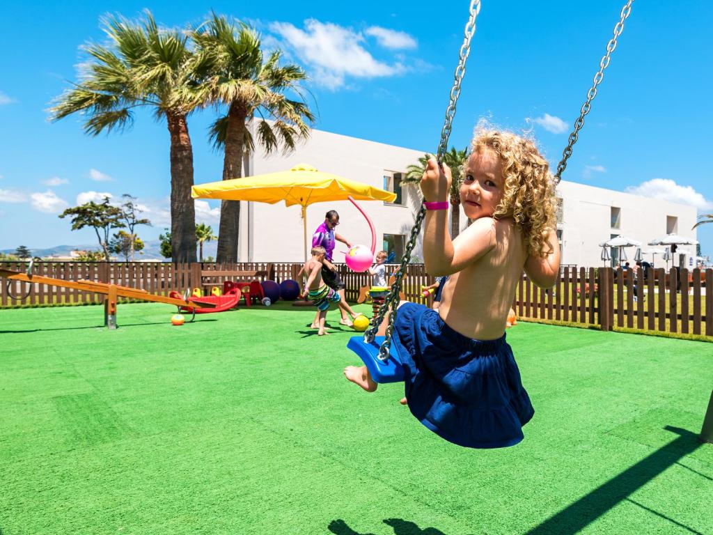 Bērns šūpolēs izbauda saulainu dienu bērnu laukumā, kam apkārt ir zaļš zāliena segums un palmas. Fons ir pilns ar rotaļlietām un citiem bērniem, kas spēlējas.