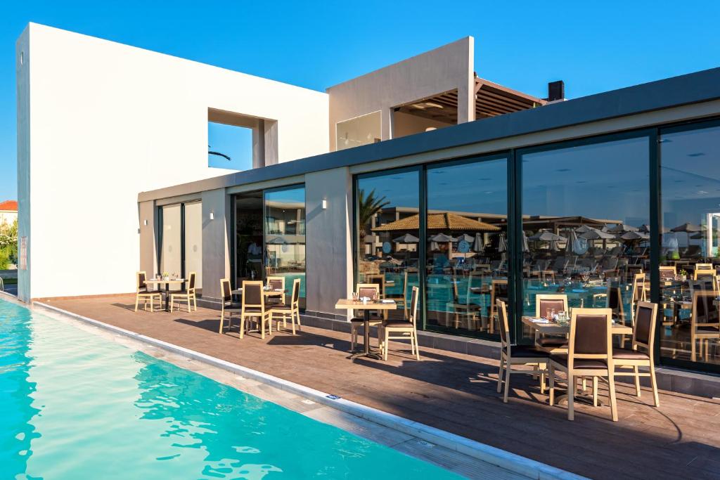 Mūsdienīga ēka ar stikla sienām, kas atrodas blakus peldbaseinam, kur pie galdiem ārpusē ir izkārti krēsli. Skats uz saulainu debesi ar palmām fona.