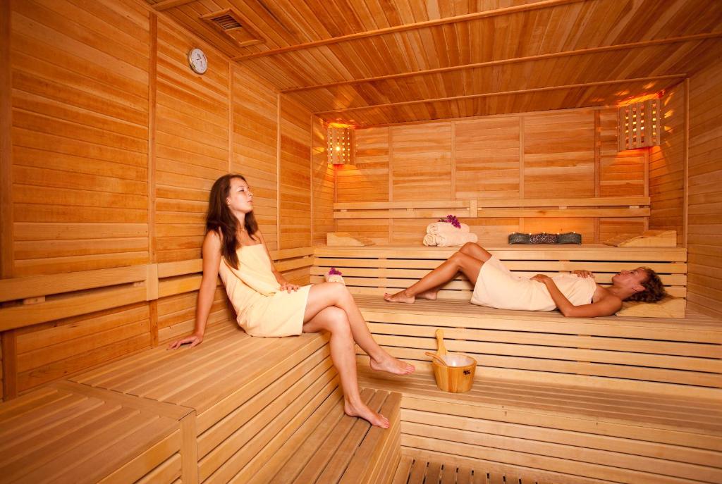 Divas sievietes relaksējas koka saunā, viena sēžot un otra guļot uz plauktiem. Saunas interjers ir koka, radot mierīgu un siltu atmosfēru.