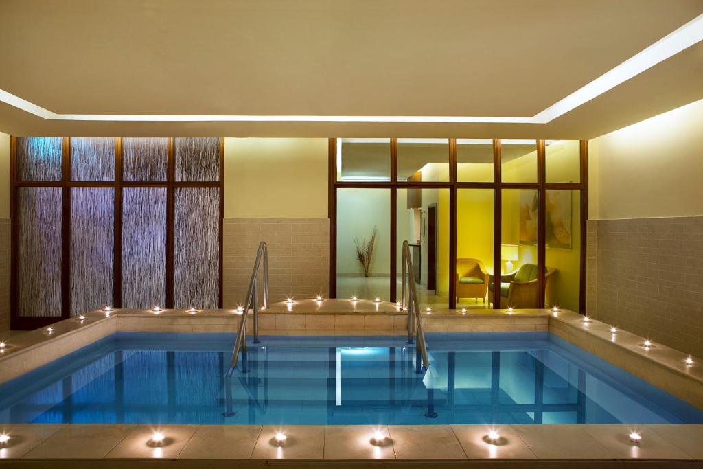 Mūsdienīgs spa ar zilu ūdeni un apgaismojumu, kas rada relaksējošu atmosfēru. Sānu daļā redzamas ērtas krēsli un lielie logi, kas ļauj ieplūst dabiskajai gaismai.