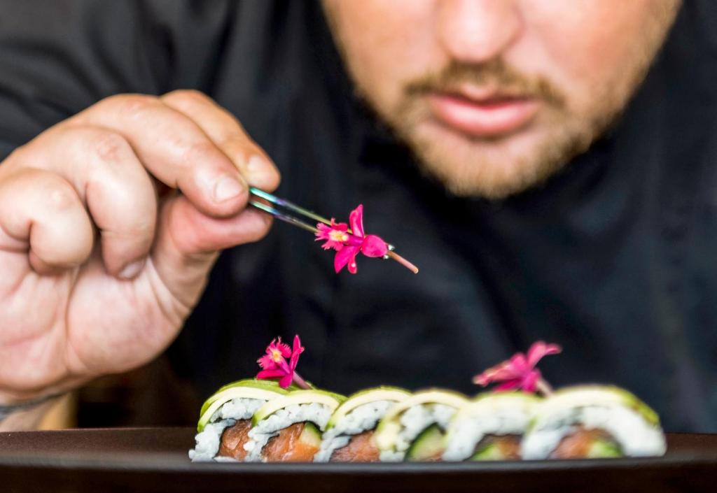 Šefpavārs dekorē suši ar krāsainām ziedu detaļām, izmantojot pinceti. Uz šķīvja redzami dažādi suši veidi, kas izskatās garšīgi un vizuāli pievilcīgi.