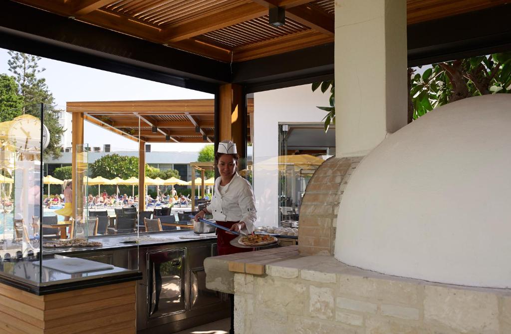 Šefpavārs pasniedz ēdienus pie āra restorāna, kur redzams peldbaseins un saulessargi. Restorāna dizains ir moderns ar koka konstrukcijām un stikla elementiem.