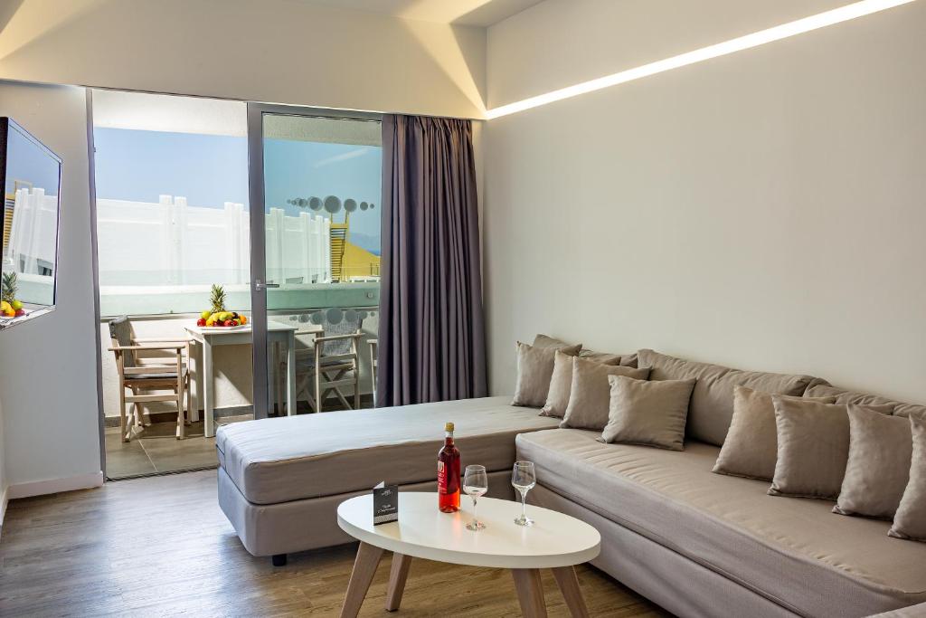 Mūsdienīga viesnīcas numura interjers ar plašu dīvānu, baltiem galdiem un piekļuvi terasei, kur redzams ēdamgaldiņš un augļu grozs.