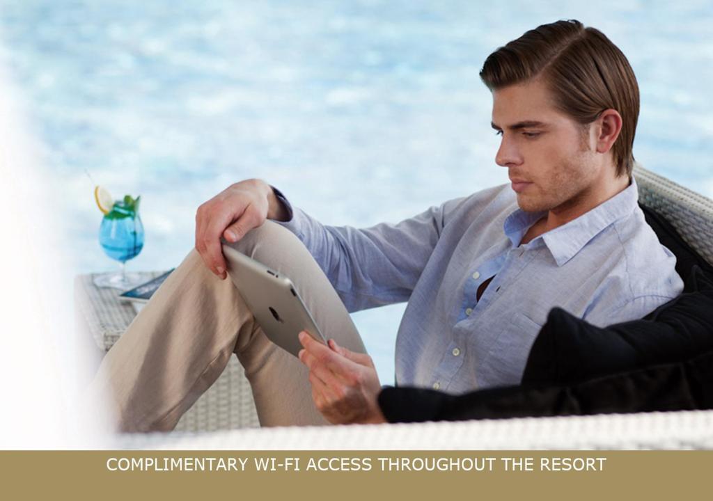 Vīrietis, kurš atpūšas pie baseina, izmantojot planšetdatoru, kamēr blakus ir dzēriens. Ir norādīts, ka viesnīcā ir pieejams bezmaksas Wi-Fi.