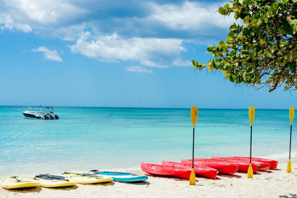 Pludmales ainava ar krāsainiem kajakiem un laivu, kas ir ūdenī. Zilais debesis un skaidrs ūdens rada mierīgu atmosfēru.
