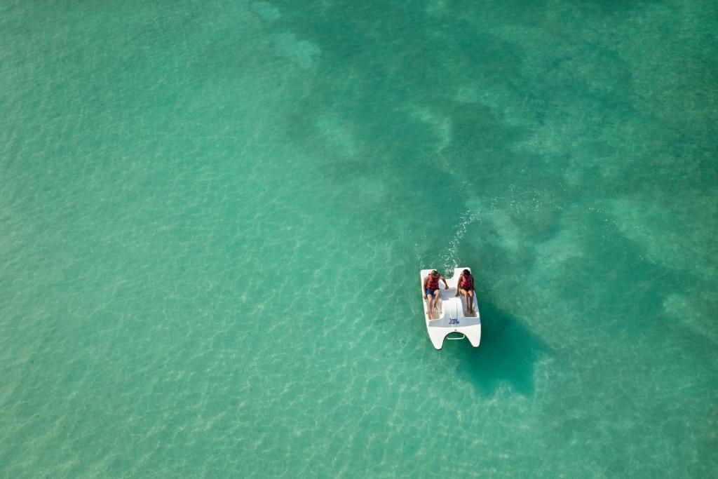 Divi cilvēki izklaidējas uz peldošā kuģī, kas peld cauri dzidro, zaļgani zilā ūdenī. Šī aina atspoguļo relaksējošu pludmales atmosfēru un vasaras priekus.