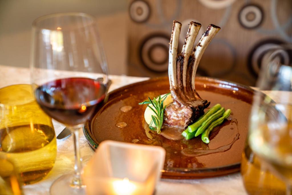 Gards un elegants ēdiens ar jēra ribām, pasniegts uz brūna šķīvja kopā ar zaļajiem pupiņām un kartupeļu biezeni, papildināts ar rosmarīnu. Fonā redzamas vīna glāzes, radot greznu atmosfēru.