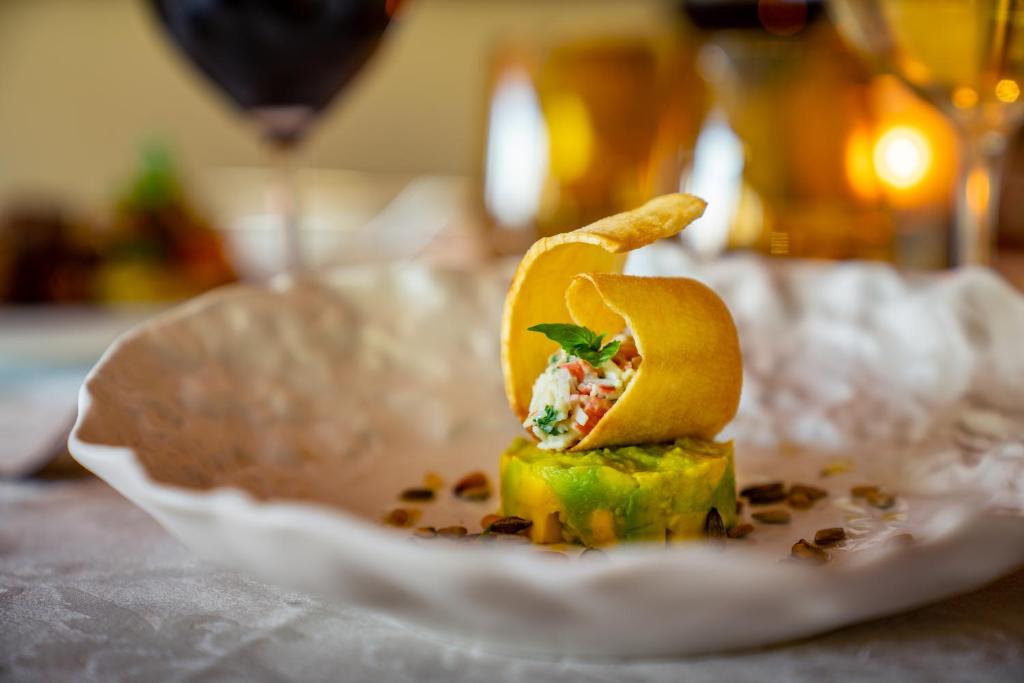 Eleganta maltīte uz dekoratīvas šķīvja, kurā redzams avokado gabals ar jūras velšu pildījumu un kraukšķīgu čipsu. Ap malām redzami rieksti un ēdieni apgaismoti ar mīkstu gaismu.