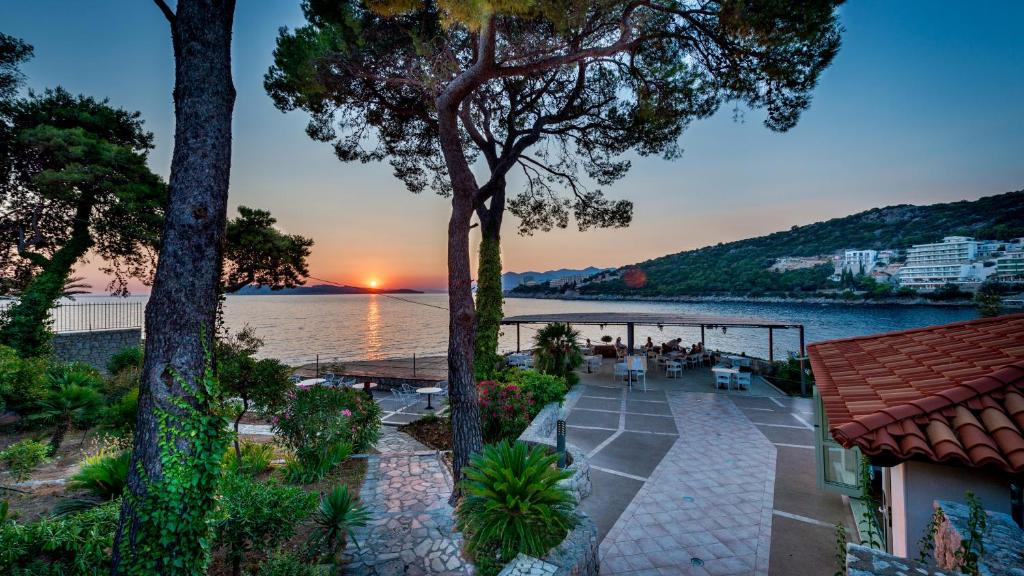 Saulrieta skats pie jūras ar koku silueti un ainaviskiem ziediem, radot mierīgu atmosfēru. Terase ar galdiem un krēsliem aicina apmeklētājus baudīt dabu.