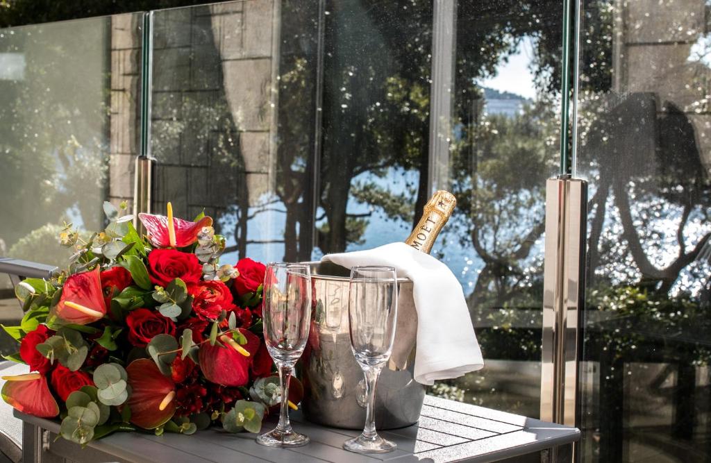 Sarkano rožu un tropisko ziedu pušķis blakus šampanieša pudelei un glāzēm, radot romantisku atmosfēru ar skatu uz ūdeni.