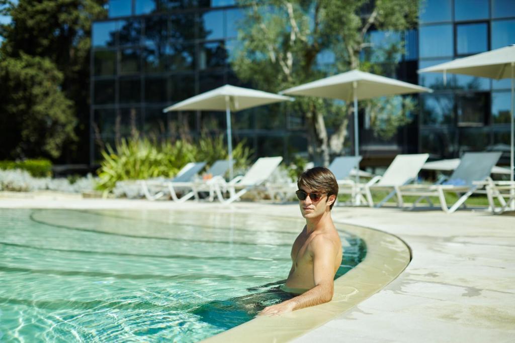 Jauns vīrietis atpūšas pie baseina, valkājot saulesbrilles, un bauda sauli. Fons ir moderns viesnīcas ēka ar baseina krēslām un sauļošanās vietām.