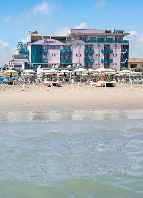 Pludmales viesnīca ar rozā fasādi un baltiem saulessargiem, kas izvietoti pie smilšu krasta. Priekšplānā redzams ūdens, kas atspoguļo debesis.