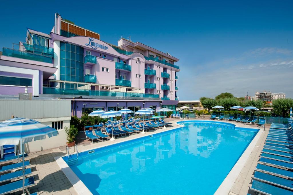 Viesnīca ar rozā fasādi un baseinu, ap kuru izvietoti sauļošanās krēsli un saulessargi. Skats uz skaidru zilo debesu fonu.