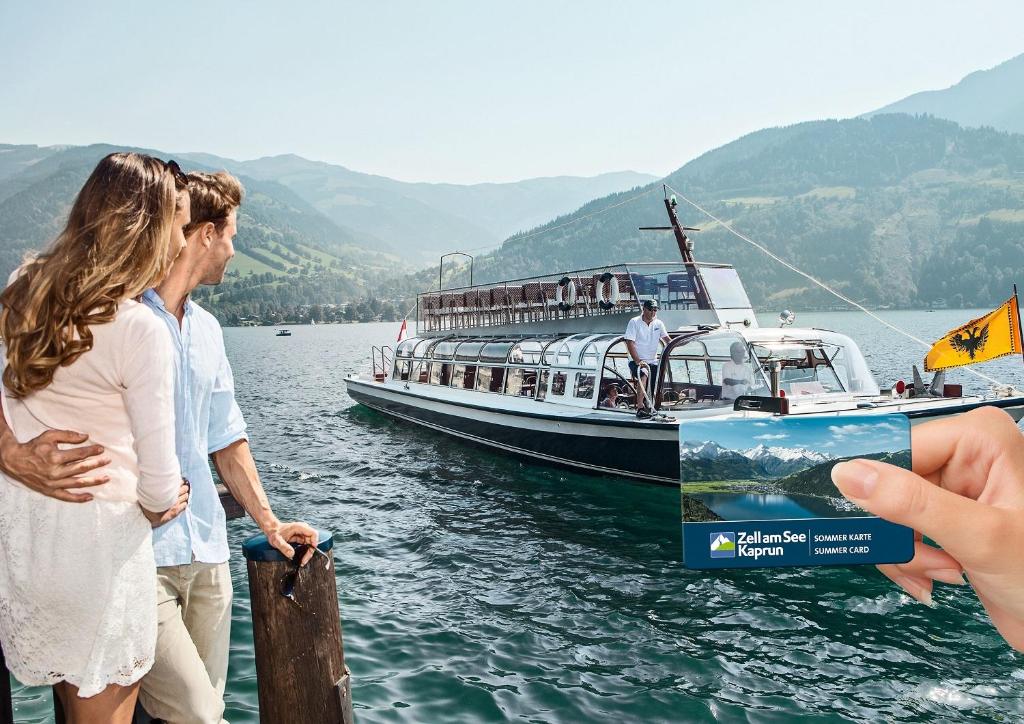 Pāris priecīgi skatās uz laivu, kas pelda gleznainā ezerā, kamēr rokā tur Zeller See Kaprun vasaras karti. Fons veido skaista kalnu ainava un saulaina debess.