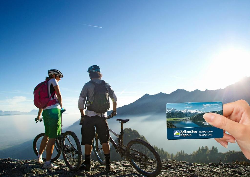 Divi kalnu riteņbraucēji, skatoties uz panorāmu, tur rokās ZellamSee Kaprun vasaras karti, fons ar kalniem un debesīm.
