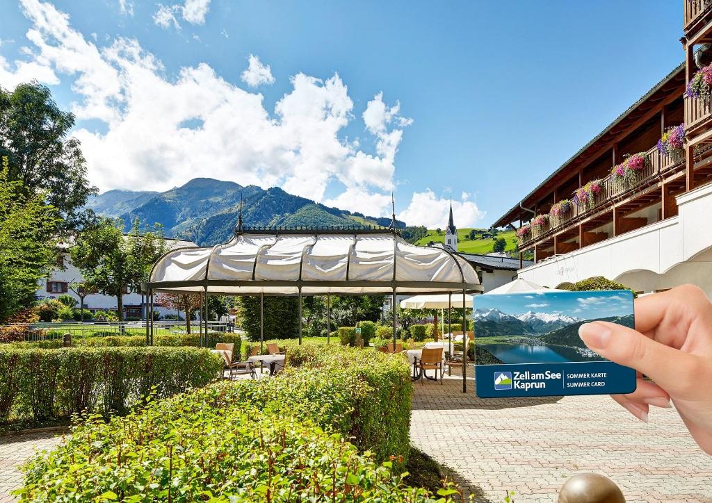 Vasaras karte "Zell am See-Kaprun" tiek turēta rokā, fons ar kalniem un viesnīcas terasi. Ideāla vieta brīvdienām dabas ielokā.