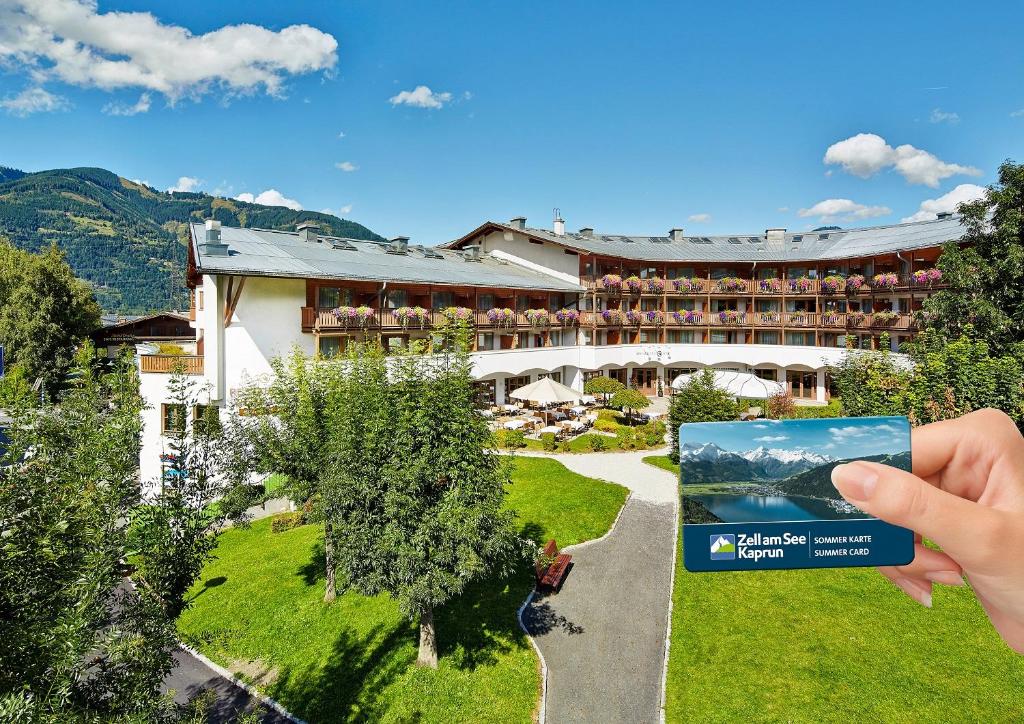 Viesnīca ar ainavisku kalnu skatu un ziedošām terasēm, kam blakus tur rokās Zell am See-Kaprun vasaras karti.