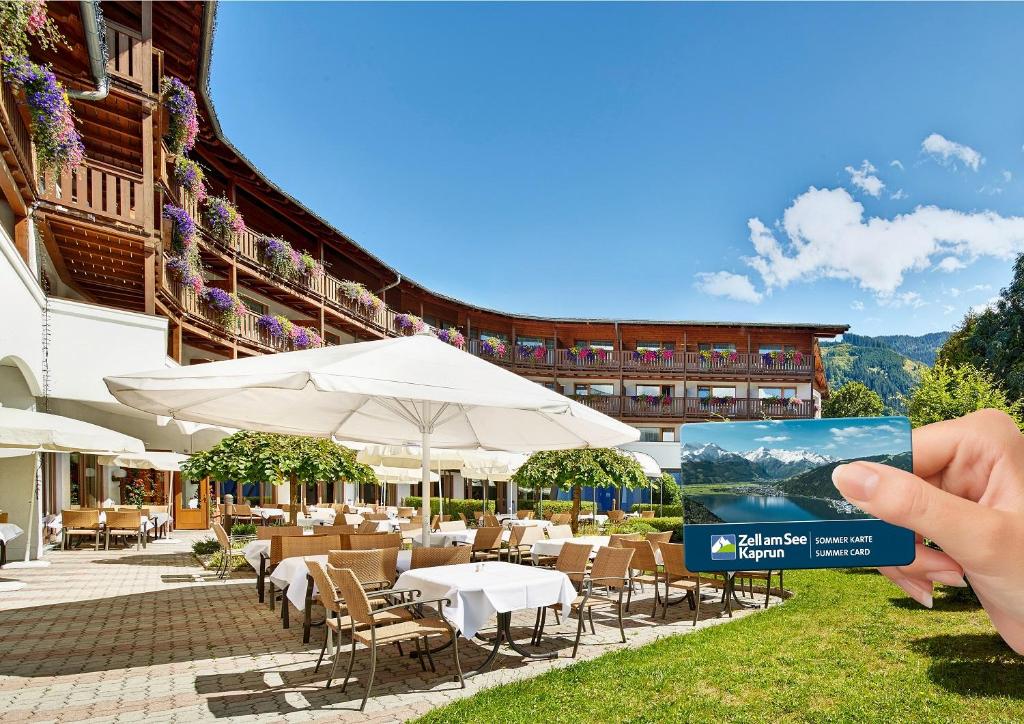 Eleganta viesnīca ar terasi un ērtām galdiem, fonā redzami kalni un zila debess. Attēlā redzama roka, kas tur "Zell am See Kaprun" vasaras karti.