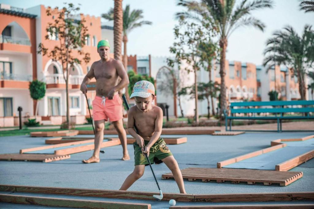 Tēls, kurā redzams tēvs un dēls, spēlējot mini golfu tropiskā vidē. Apkārt ir palmām apstādīta teritorija un viesnīcas ēkas.
