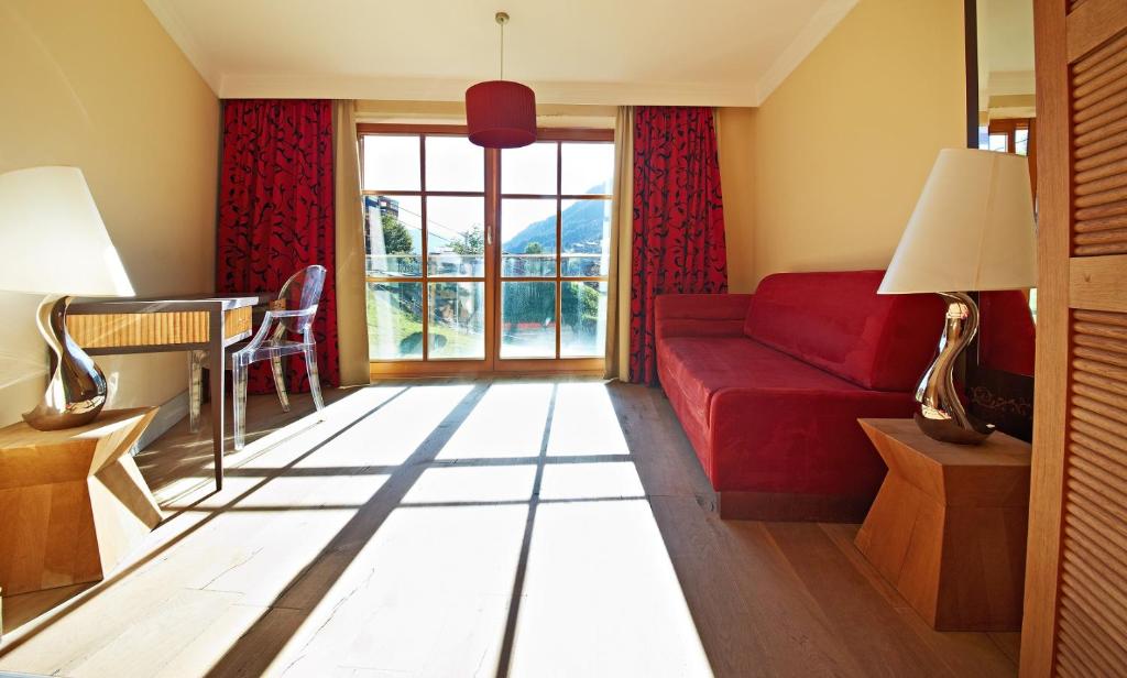 Mūsdienīgs viesnīcas numurs ar sarkanu dīvānu, koka grīdu un lieliem logiem, caur kuriem ieplūst dabiska gaisma. Interjers ir elegants, ar modernām mēbelēm un siltām krāsām.