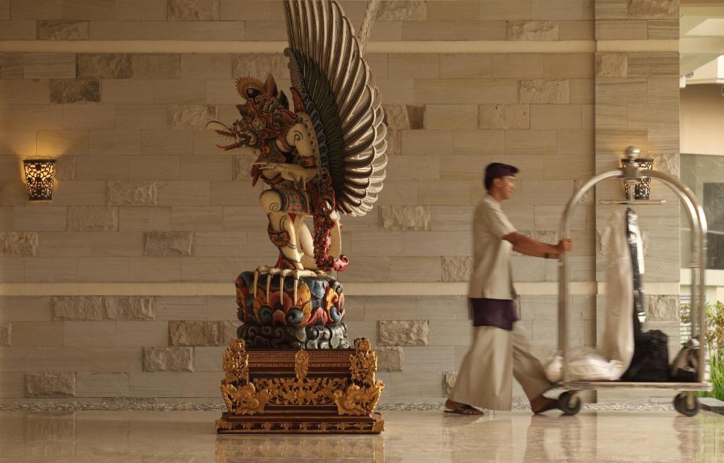 Lieliska viesnīcas recepcija ar tradicionālu statuju un darbinieku, kurš stumj bagāžas ratiņus. Iekšējā apdare apvieno mūsdienīgu un tradicionālo stilu.