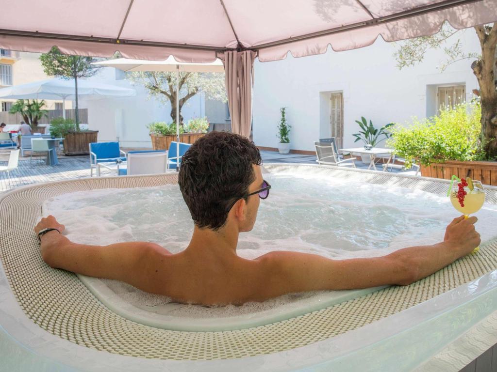 Vīrietis relaksējas džakuzi, turot kokteili rokās, kamēr apkārt ir zaļumi un atpūtas zonas. Ideāla atmosfēra brīvā laika pavadīšanai.