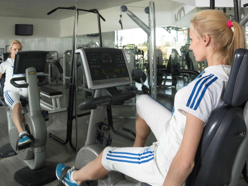 Sieviete vingro sporta zālē uz velotrenažiera, skatoties spogulī. Aiz viņas redzami citi trenažieri un modernas sporta iekārtas.