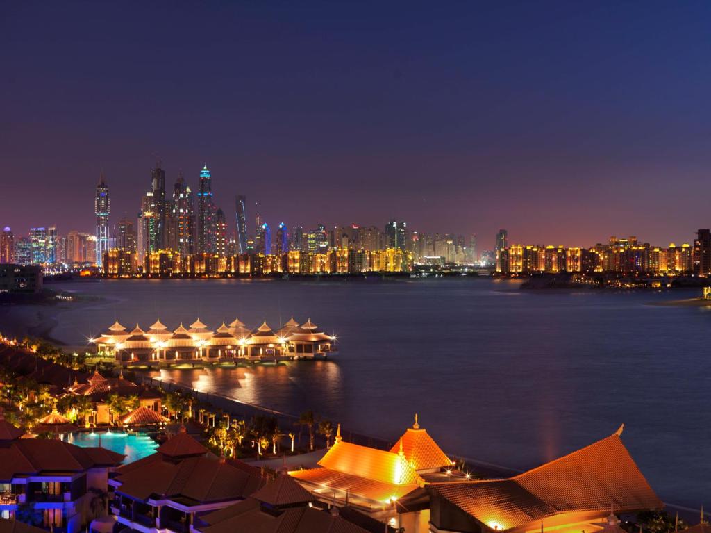 Nakts skats uz Dubaiju, kur izceļas mūsdienu debesskrāpji un gaismas atspīd ūdenī. Priekšplānā redzamas elegantas ēkas ar sarkaniem jumtiem un krāšņs ainavas apgaismojums.