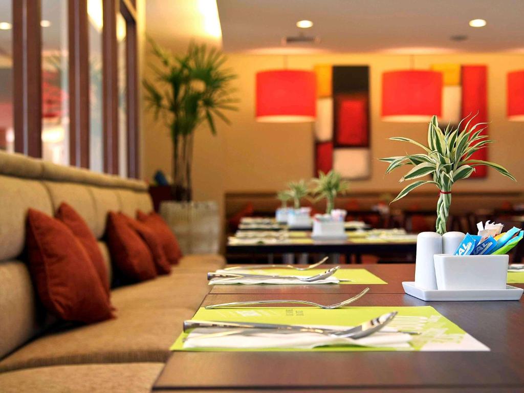 Restorāna interjers ar elegantām galda klāšanas detaļām, sarkaniem spilveniem un modernu apgaismojumu. Zaļi galdauti un dekoratīvi augi piešķir telpai svaigumu.