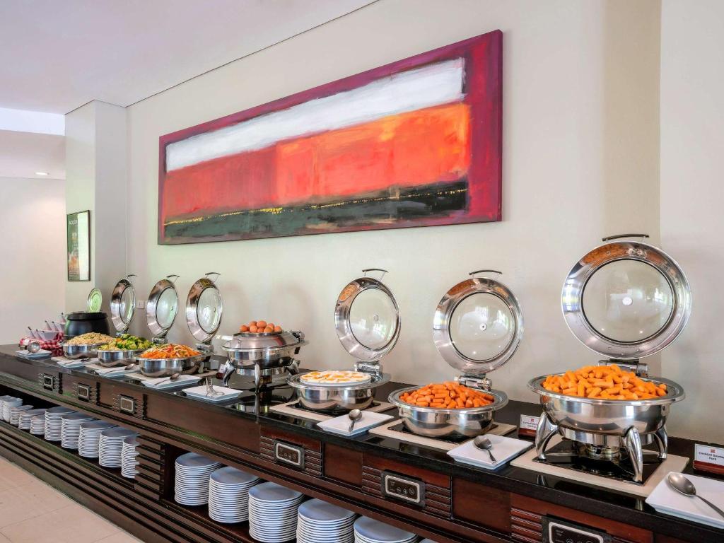 Bagātīgs brokastu bufete ar dažādiem ēdieniem, tostarp augļiem un dārzeņiem, izvietota pie elegantas koka galda. Sienā redzama moderna mākslas glezna.