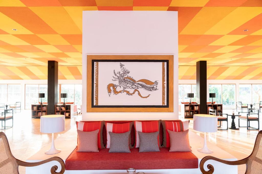 Mūsdienīgs interjers ar spilgtu oranžu un dzeltenu griestu dizainu, centrā redzama mākslas darbu kompozīcija. Komfortabli sēdekļi un apgaismojums rada patīkamu atmosfēru.