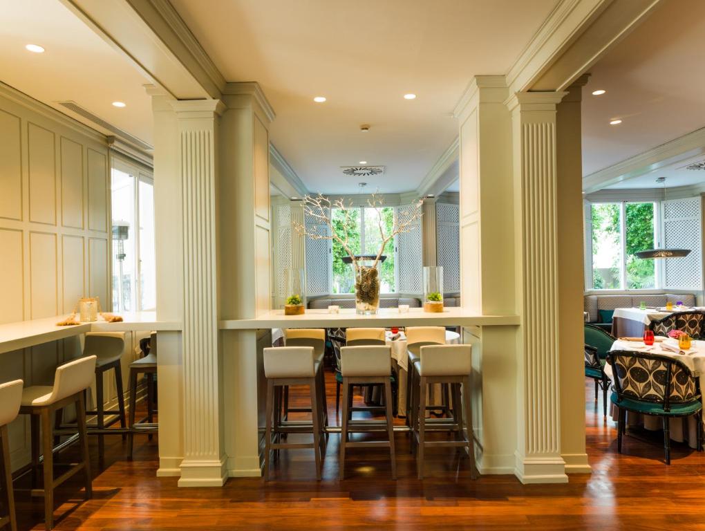 Elegants restorāns ar mūsdienīgu interjeru, kurā ir gaišas krāsas un koka grīda, radot patīkamu atmosfēru maltītēm. Stilīgs bārs ir redzams priekšplānā, bet fonā ir ērtas sēdvietas un dabiska apgaismojuma avoti.