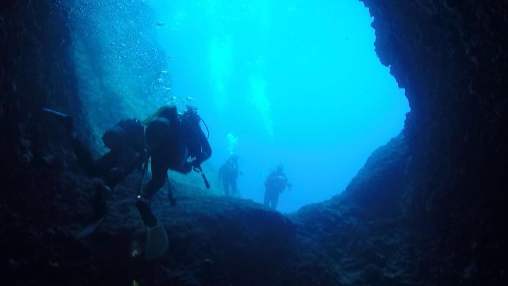 Apakšūdens skats ar nirējiem, kas pēta klinšu veidojumus zem ūdens, ar spilgti zilo ūdeni fonā.