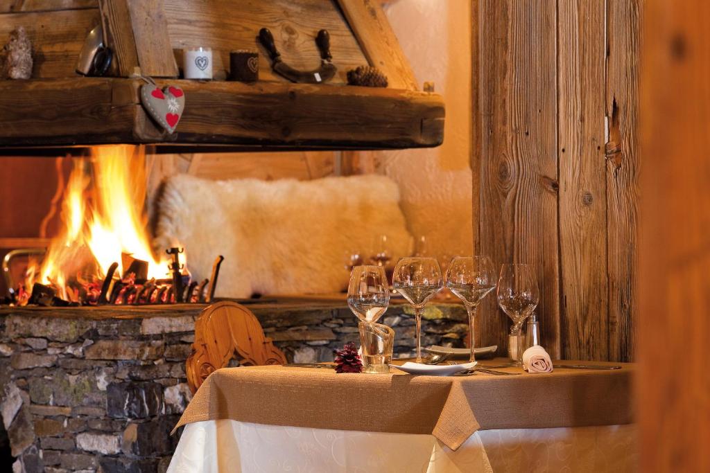 Romantiska ēdamzona ar elegantu galdu, ko rotā glāzes un ziedu kompozīcija, kamēr kamīnā deg uguns, radot siltu un mājīgu atmosfēru.