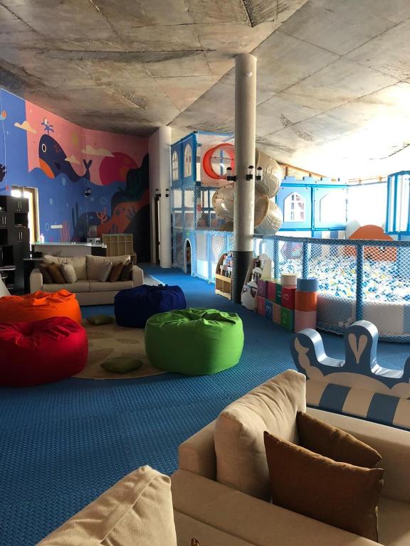 Krāsaina bērnu rotaļu telpa ar mīkstiem sēdvietām un interaktīviem elementiem, piemēram, krāsainiem kubiem un rotaļu laukumu. Telpas sienas rotā jūras tematika, radot draudzīgu un jautru atmosfēru.